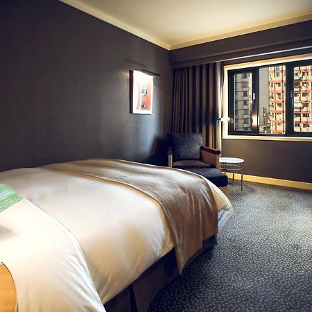 Ambassador Hotel Kaohsiung Exterior photo The photo shows a hotel room featuring a large bed with a light-colored duvet. The walls are painted in a dark hue, creating a cozy atmosphere. There is a chair and a small side table near the window, which is covered by curtains. Through the window,