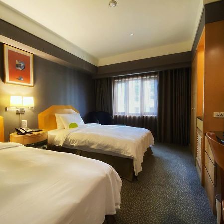 Ambassador Hotel Kaohsiung Exterior photo The photo shows a hotel room with two beds, each made up with white linens. The walls are painted in dark colors, while the bedding and furniture are lighter in tone. There is a nightstand between the beds with a lamp and telephone on it. A window wi