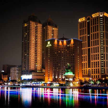 Ambassador Hotel Kaohsiung Exterior photo The photo depicts a vibrant nighttime cityscape featuring several tall buildings illuminated by colorful lights. Prominent structures include a large hotel and other high-rise buildings with illuminated facades. In front of these buildings, there app