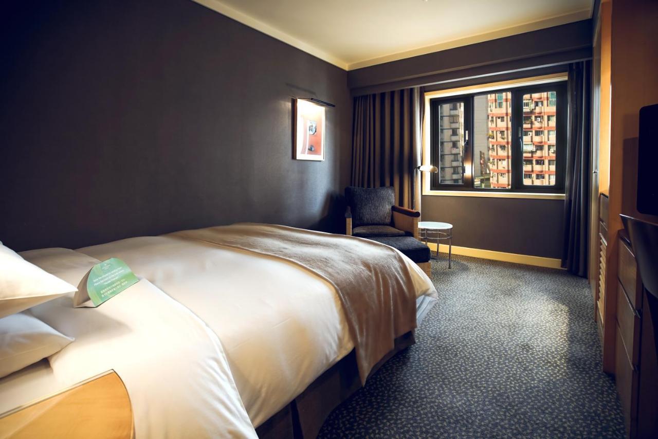 Ambassador Hotel Kaohsiung Exterior photo The photo shows a hotel room featuring a large bed with a light-colored duvet. The walls are painted in a dark hue, creating a cozy atmosphere. There is a chair and a small side table near the window, which is covered by curtains. Through the window,