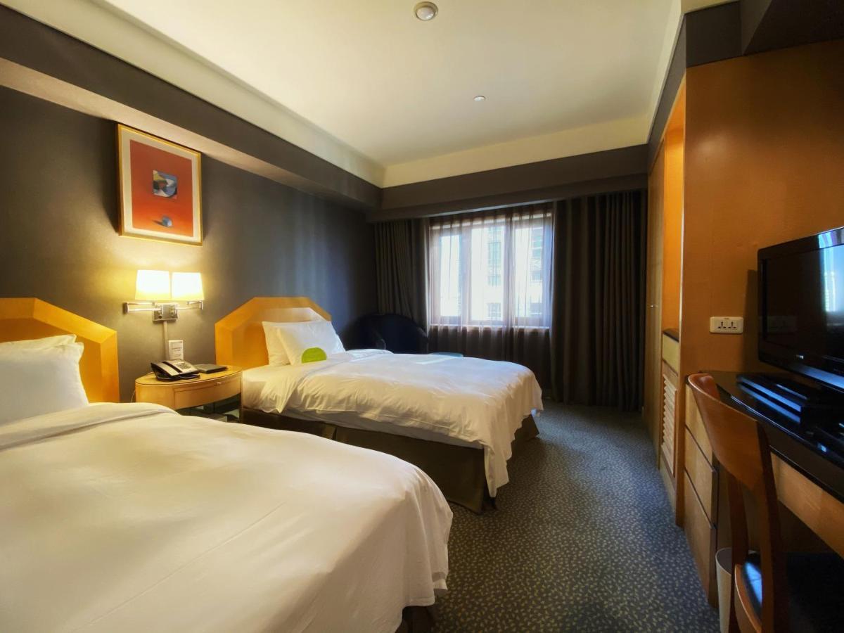 Ambassador Hotel Kaohsiung Exterior photo The photo shows a hotel room with two beds, each made up with white linens. The walls are painted in dark colors, while the bedding and furniture are lighter in tone. There is a nightstand between the beds with a lamp and telephone on it. A window wi