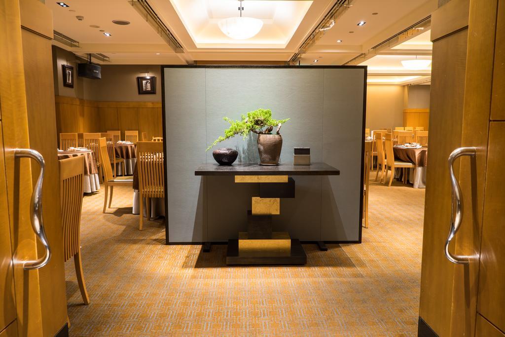 Ambassador Hotel Kaohsiung Exterior photo The photo shows the interior of a restaurant. In the foreground, there is a decorative table with a minimalist design, featuring a vase with greenery. The background reveals a spacious dining area with wooden chairs and tables, creating a warm and in