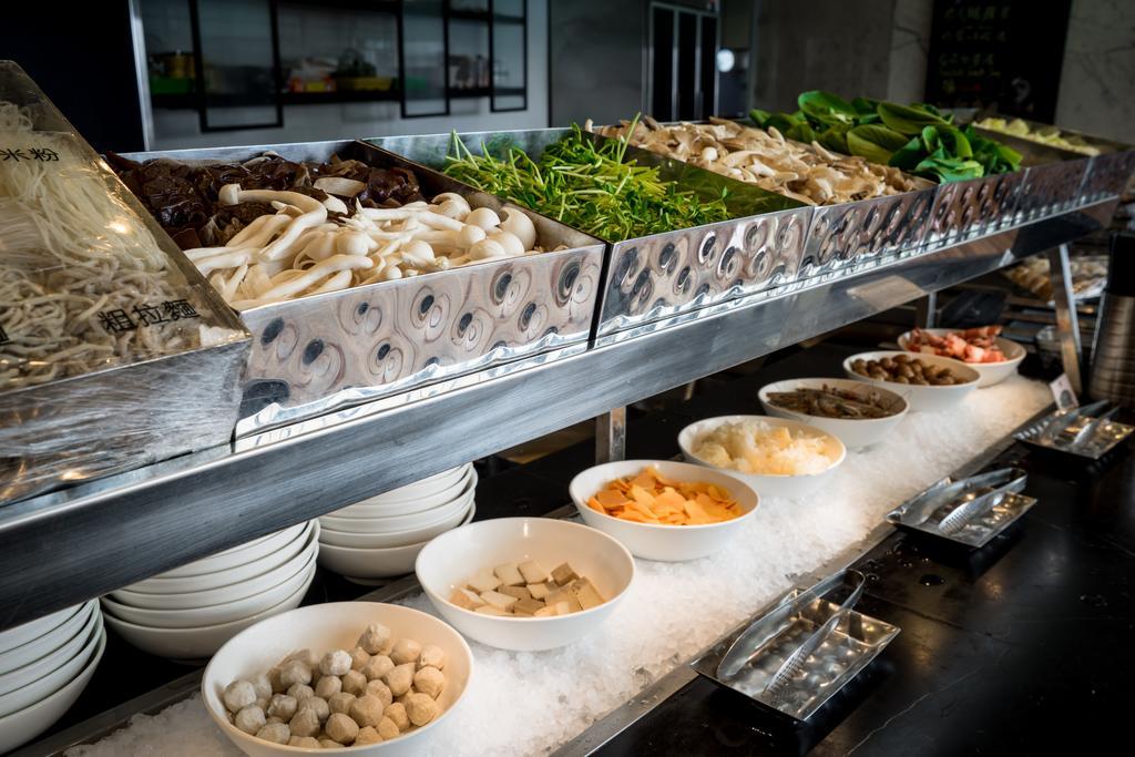 Ambassador Hotel Kaohsiung Exterior photo The photo shows a buffet or display of various food items, likely from an Asian cuisine setting. There are several metal trays filled with a variety of fresh vegetables, including greens and mushrooms. Below these trays, there are white bowls contain
