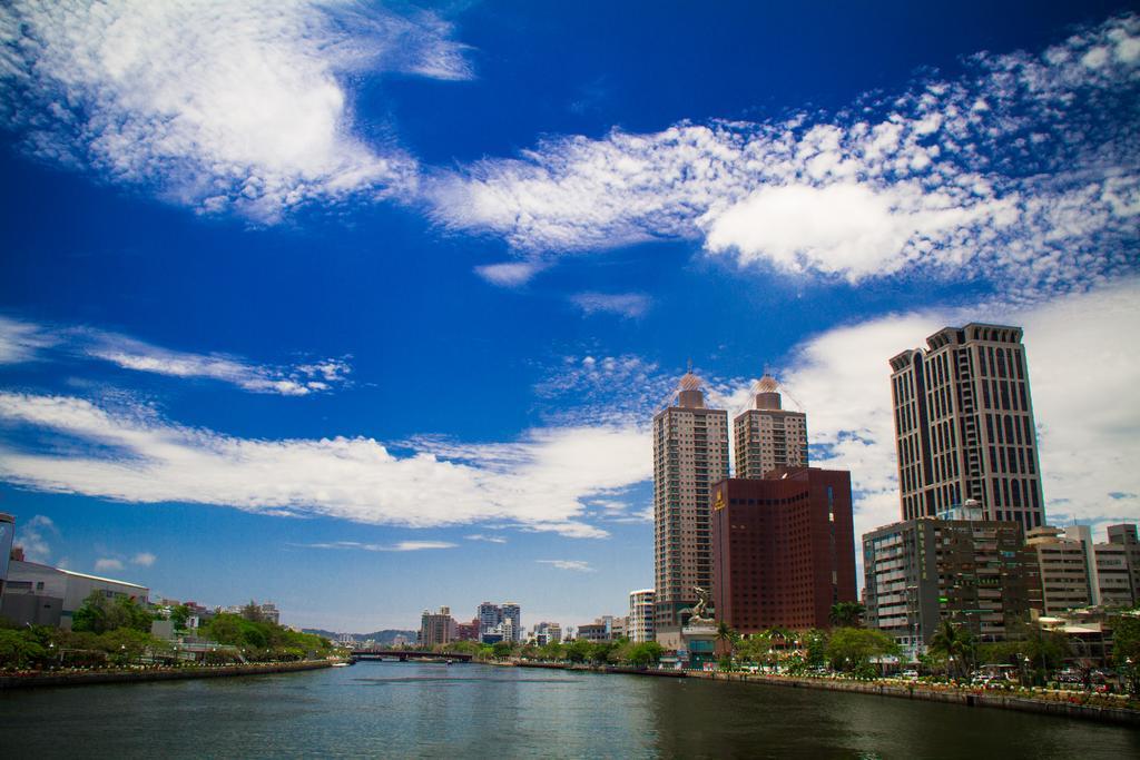 Ambassador Hotel Kaohsiung Exterior photo The photo showcases a scenic view of a river flanked by modern skyscrapers. The buildings have a mix of architectural styles, including tall, glass-fronted structures and a reddish-brown building. In the background, a clear blue sky features fluffy w