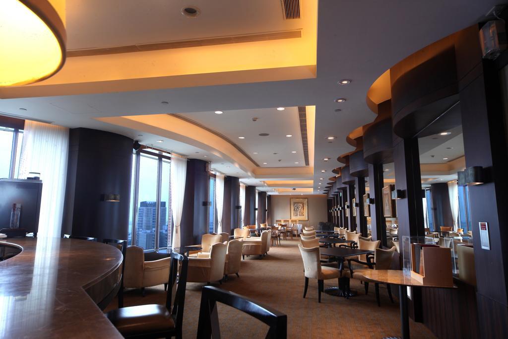 Ambassador Hotel Kaohsiung Exterior photo The photo shows the interior of a modern restaurant or lounge. The space features elegant seating with cream-colored chairs and wooden tables, arranged in a spacious layout. Soft, warm lighting highlights the architectural details of the ceiling and 