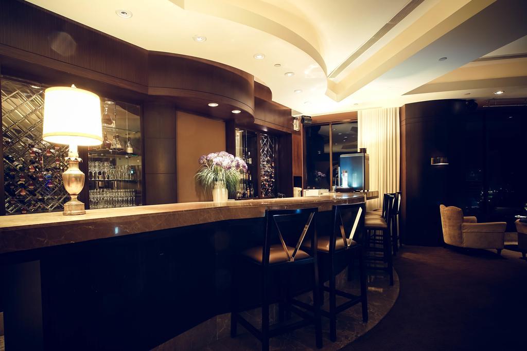 Ambassador Hotel Kaohsiung Exterior photo The photo shows a modern and stylish bar area. The bar is made of a smooth stone surface and features a backlit shelf displaying various bottles of liquor. There is a lamp on the bar providing warm lighting, and a vase with flowers adds a decorative 
