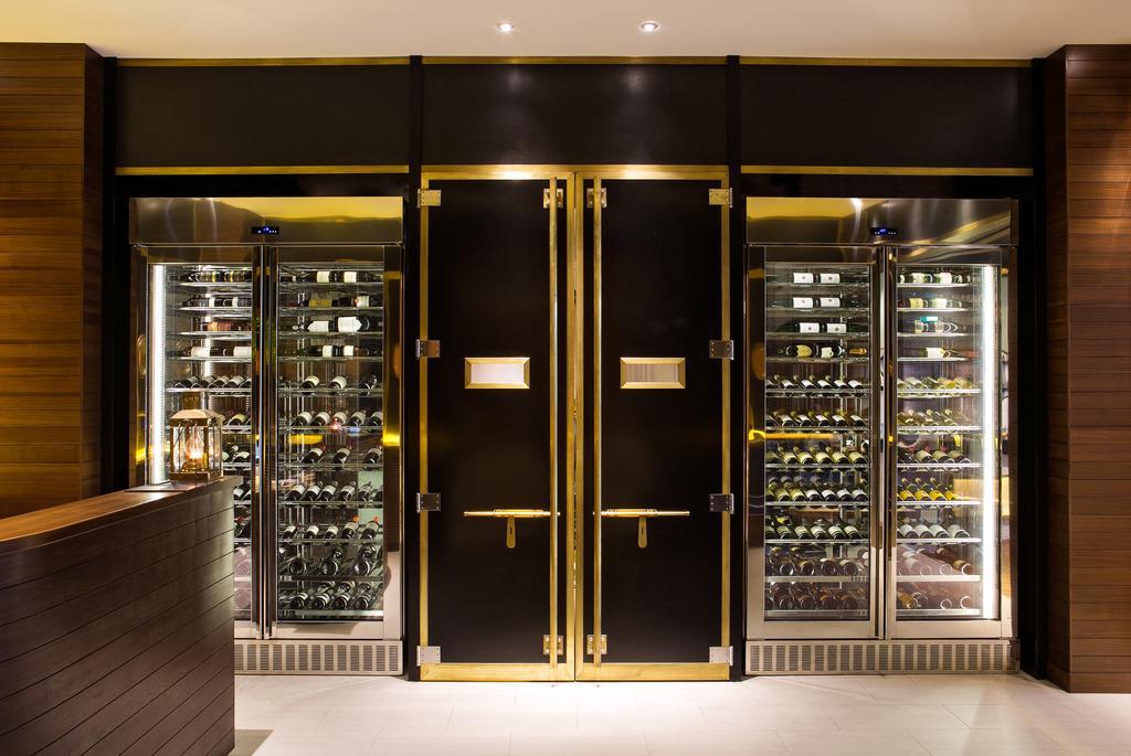 Ambassador Hotel Kaohsiung Exterior photo The photo shows a stylish, modern display area, likely in a restaurant or wine bar. It features large black double doors with gold trim at the center, flanked by glass refrigeration units on either side. The glass units are filled with various bottle