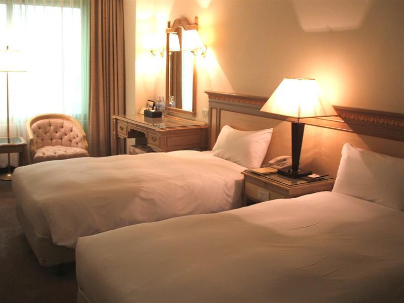 Ambassador Hotel Kaohsiung Exterior photo The photo shows a cozy hotel room with two single beds, each covered with white bedding. There is a lamp on a nightstand between the beds. A wooden dresser with a mirror is against the wall, and a comfortable armchair is placed nearby. Soft lighting 