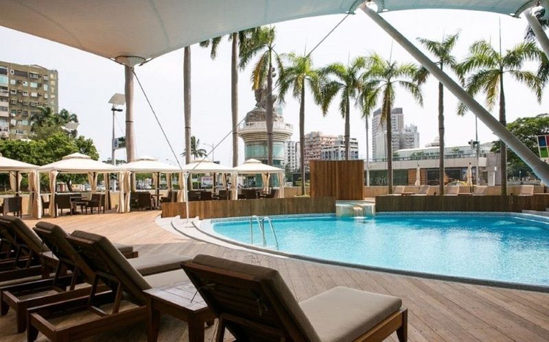 Ambassador Hotel Kaohsiung Exterior photo The photo shows a modern outdoor pool area. It features a clear blue swimming pool surrounded by wooden decking. There are comfortable lounge chairs positioned near the pool, and several large umbrellas provide shade. In the background, tall palm tre