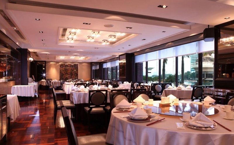 Ambassador Hotel Kaohsiung Exterior photo The photo shows an elegant dining room with a modern and upscale design. The space features round tables covered with white tablecloths, each set with neatly arranged dinnerware, including folded napkins. The floor is made of polished wood, enhancing
