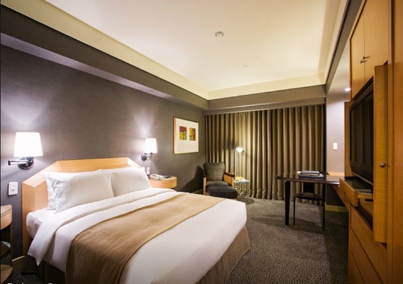 Ambassador Hotel Kaohsiung Exterior photo The photo shows a modern hotel room featuring a neatly made bed with white linens and a beige bedspread. There are bedside lamps on either side of the bed. One corner of the room includes a comfortable armchair, and adjacent to it is a small table. T
