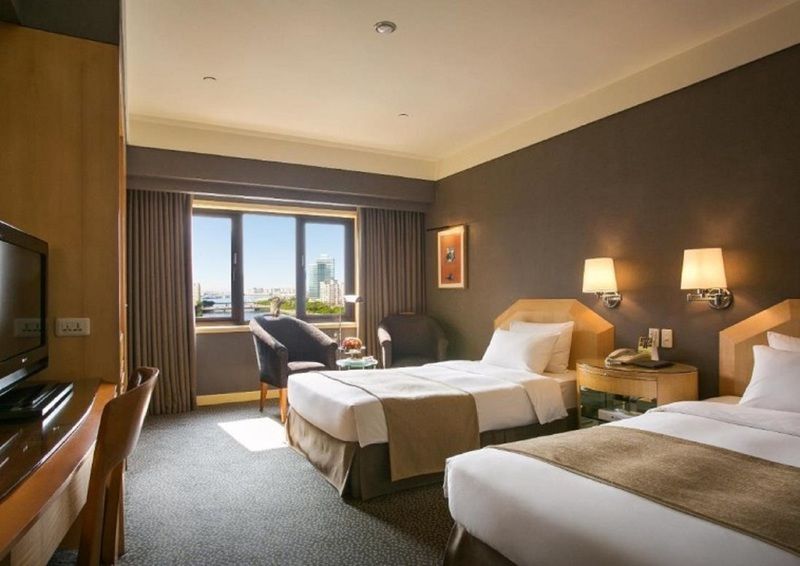Ambassador Hotel Kaohsiung Exterior photo The photo shows a hotel room featuring two twin beds with beige and brown bedding. The room has a modern design with a dark accent wall and light-colored furniture. There are bedside lamps on each side of the beds, and a comfortable armchair is posit