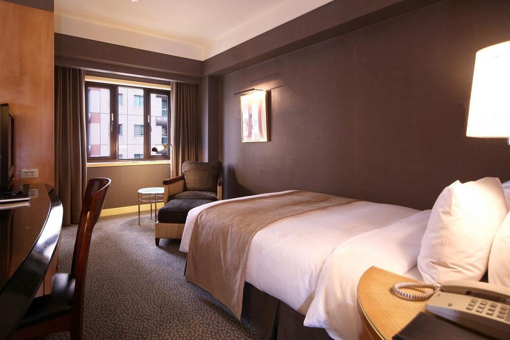 Ambassador Hotel Kaohsiung Exterior photo The photo shows a hotel room featuring a neatly made single bed with a light-colored blanket. There is a comfortable chair in the corner alongside a small round table. A large window allows natural light to enter the room, and the walls are painted i