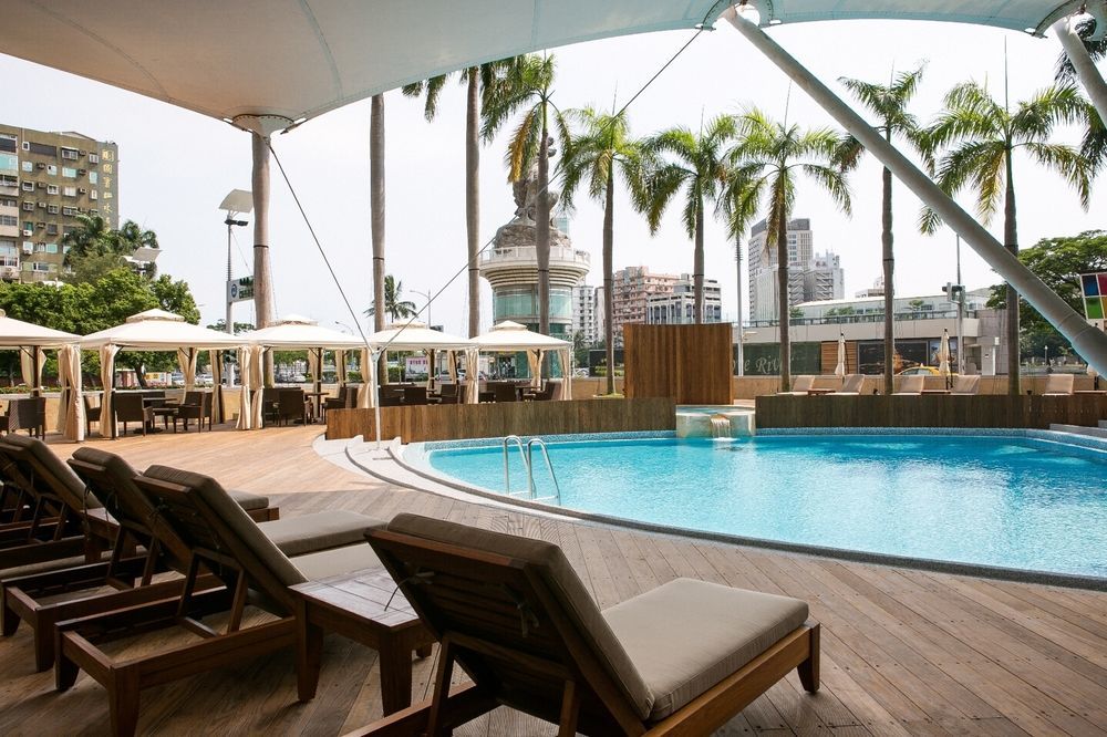 Ambassador Hotel Kaohsiung Exterior photo The photo shows a resort-style pool area. In the foreground, there are several wooden lounge chairs arranged around a sparkling blue pool. The pool has a gentle slope and is surrounded by wooden decking. Behind the pool, tall palm trees add to the tr