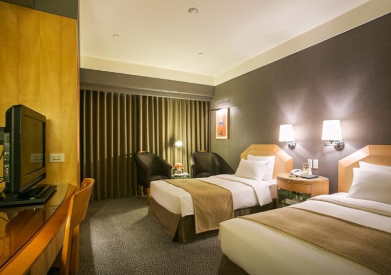 Ambassador Hotel Kaohsiung Exterior photo The photo shows a hotel room featuring two single beds with light-colored bedding. The walls are painted in a muted color, and there are two wall-mounted lamps above each bed. A wooden desk and chair are positioned against one wall, accompanied by a 