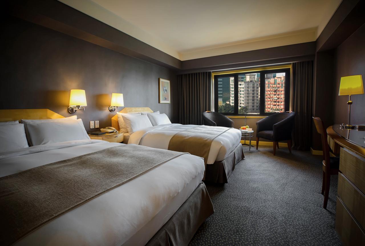 Ambassador Hotel Kaohsiung Exterior photo The photograph shows a hotel room featuring two double beds with white linens and light brown accents. The walls are painted a dark color, and there are bedside lamps beside each bed. A window offers a view of the outside, and there are two chairs po
