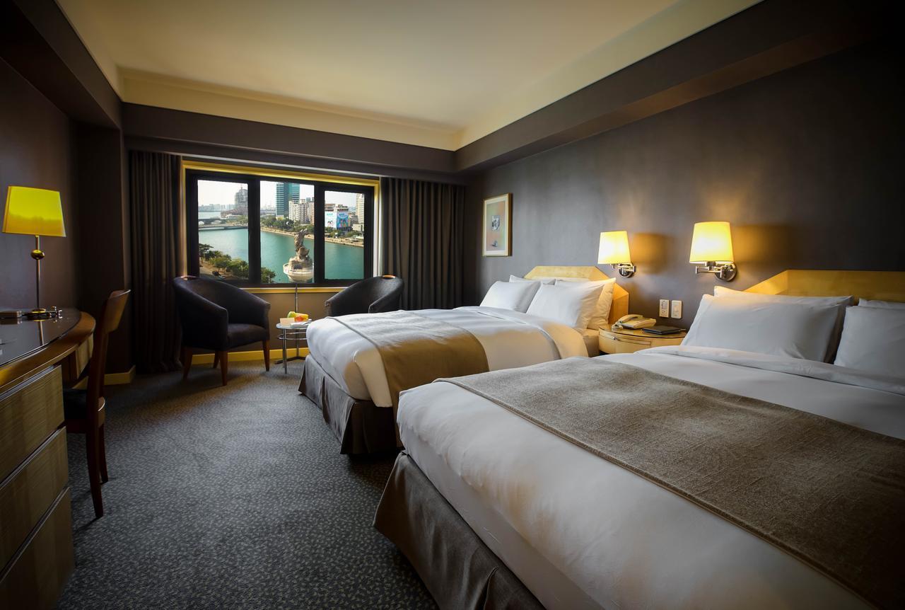 Ambassador Hotel Kaohsiung Exterior photo The photo shows a hotel room featuring two large beds with white linens and beige accents. The room has a modern design with dark walls and plush carpeting. There are lamps on each side of the beds, providing warm lighting. A large window offers a vi