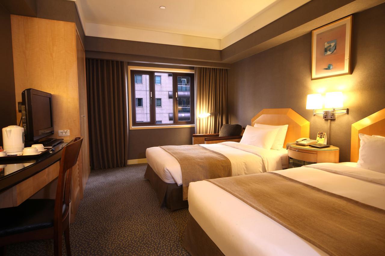 Ambassador Hotel Kaohsiung Exterior photo The photo shows a hotel room featuring two twin beds with neutral-colored bed linens. The room has a modern design, with warm lighting and wooden furnishings. There's a desk and chair positioned against one wall, along with a television. A large wind