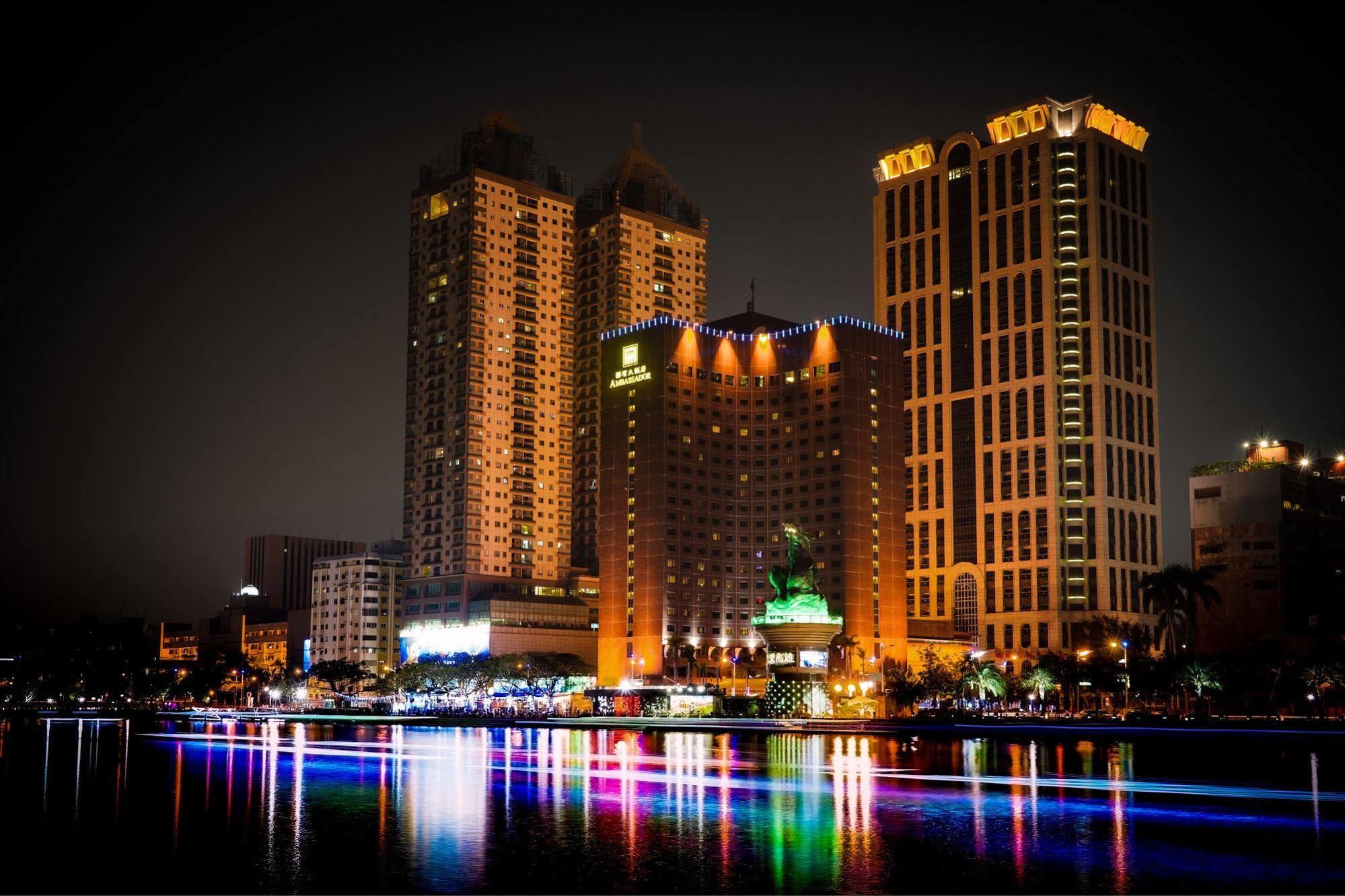 Ambassador Hotel Kaohsiung Exterior photo The photo depicts a vibrant nighttime cityscape featuring several tall buildings illuminated by colorful lights. Prominent structures include a large hotel and other high-rise buildings with illuminated facades. In front of these buildings, there app