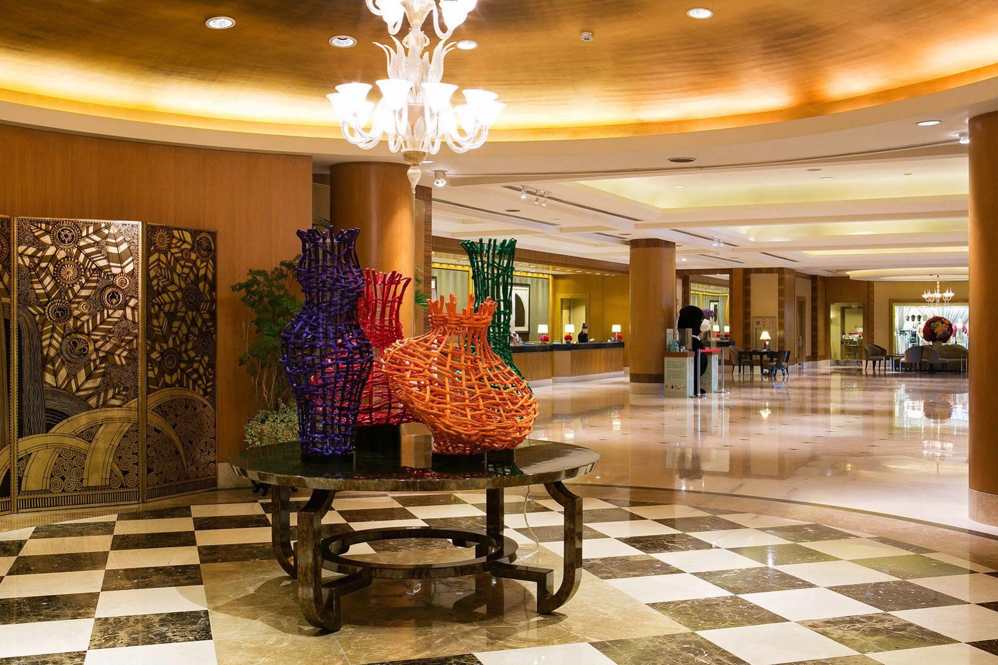 Ambassador Hotel Kaohsiung Exterior photo The photo shows the interior of a hotel lobby featuring an elegantly designed space. In the foreground, there is a decorative table with colorful woven vases, predominantly in purple and orange. These vases add a vibrant touch to the setting. The flo