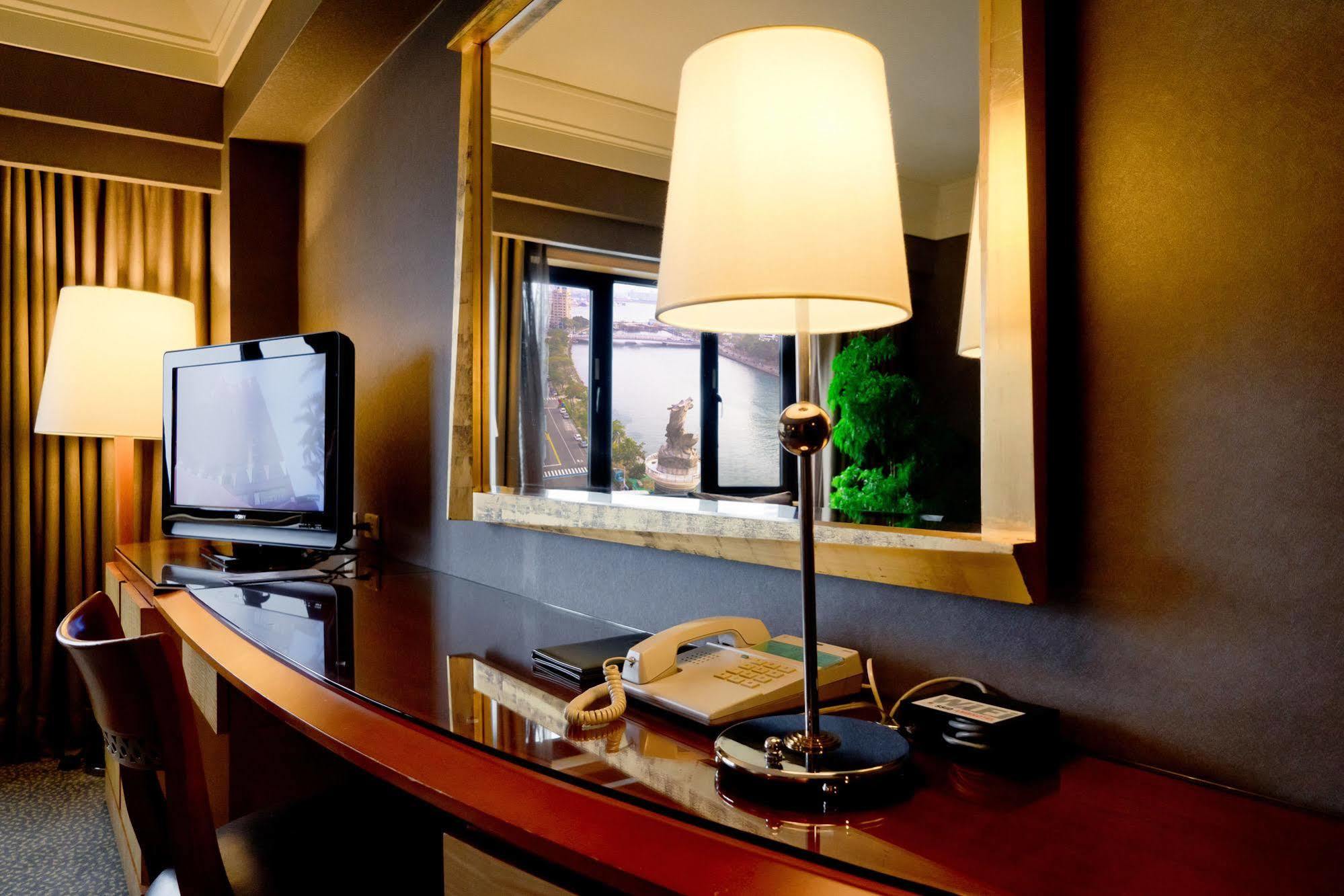 Ambassador Hotel Kaohsiung Exterior photo The photo depicts a hotel room interior featuring a sleek wooden desk with a curved design. On the desk, there is a modern lamp with a white shade, a telephone, and a television set to the left. A mirror hangs above the desk, reflecting light and the