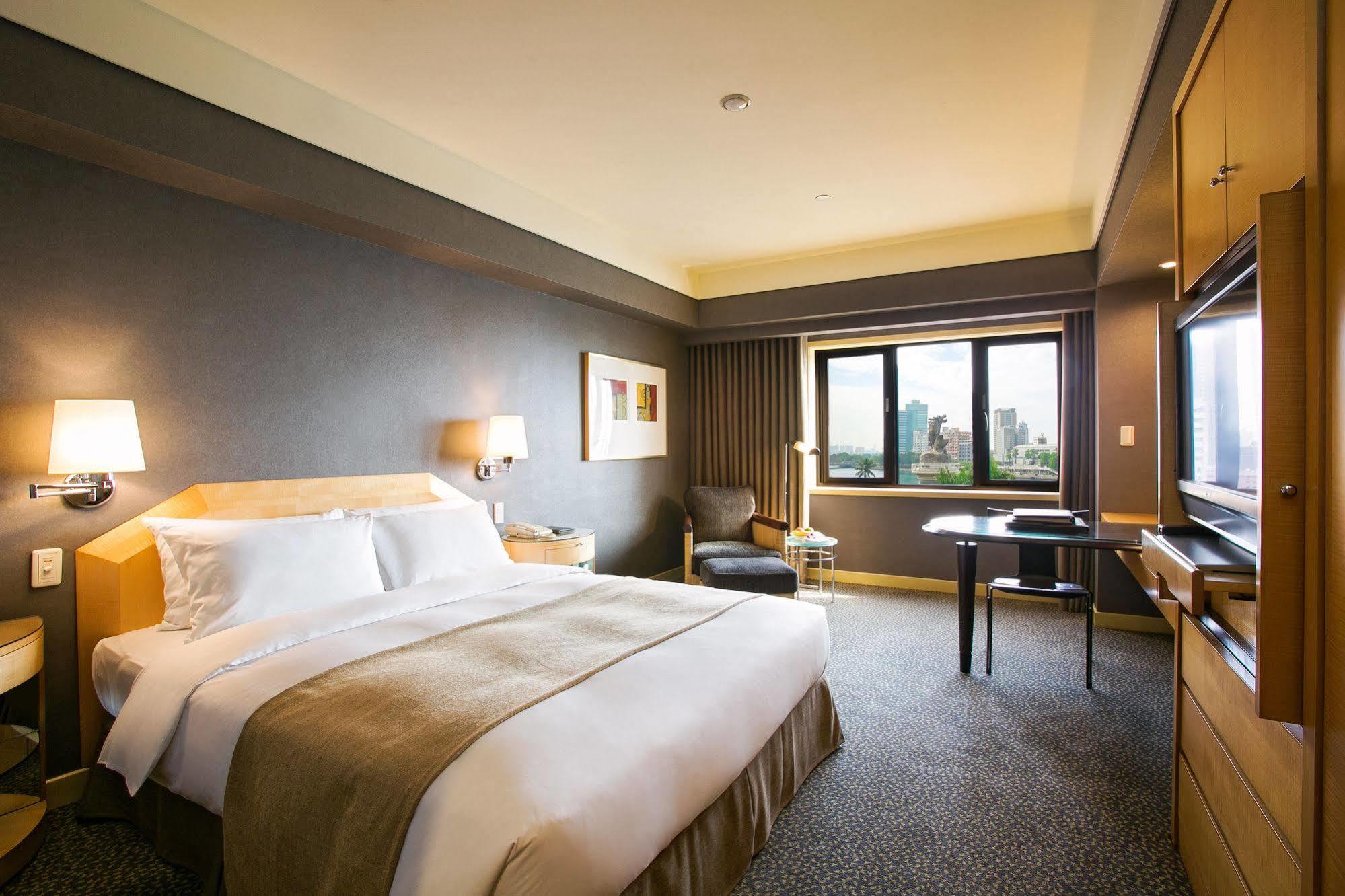 Ambassador Hotel Kaohsiung Exterior photo The photo shows a hotel room that features a large bed with crisp white linens and a beige throw blanket. There are bedside lamps on either side of the bed. A comfortable chair is positioned near a window with light drapes, and a small desk with a ch