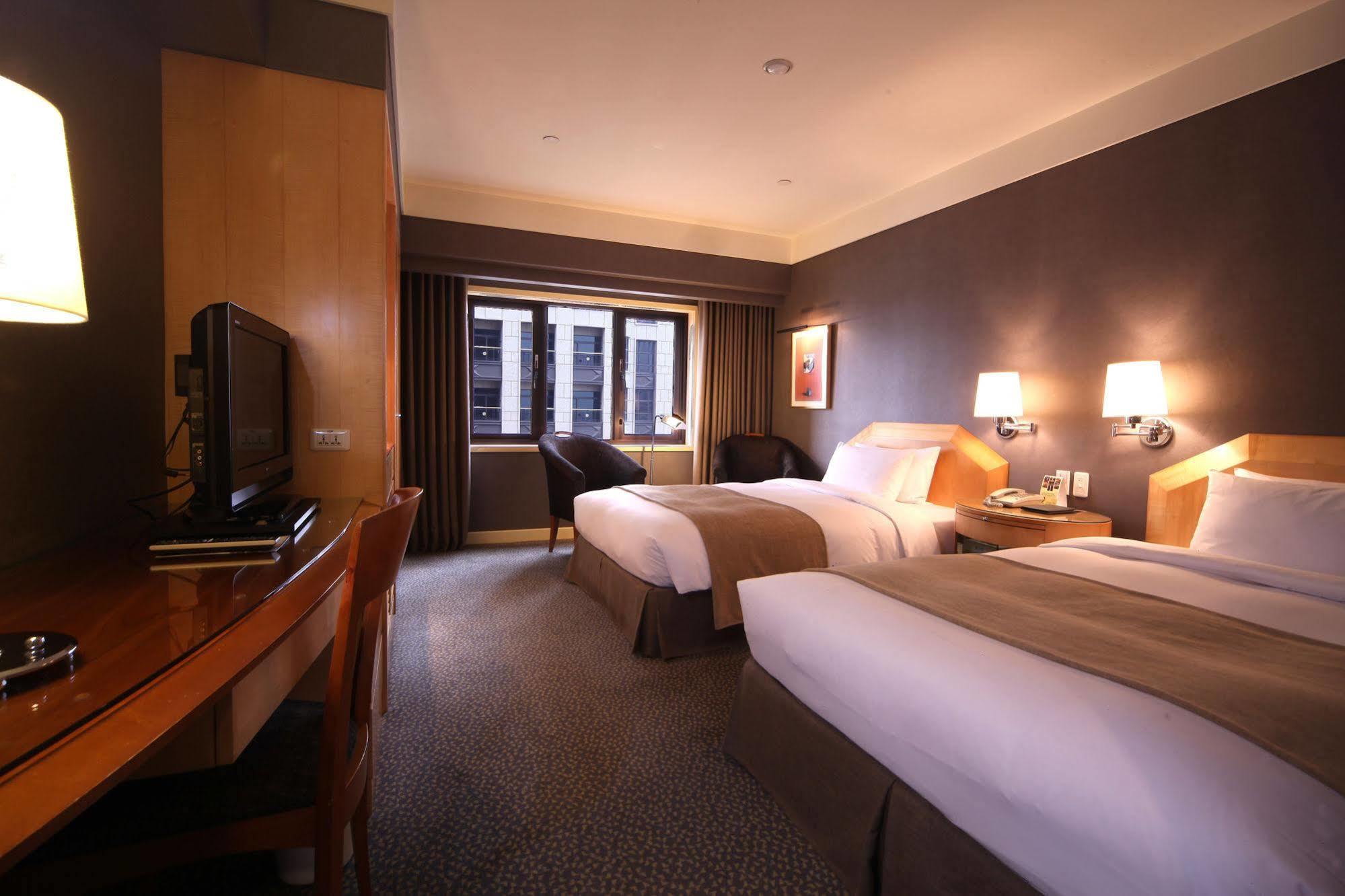 Ambassador Hotel Kaohsiung Exterior photo The photo shows a hotel room featuring two twin beds, each with light-colored bedding. There are bedside lamps on each side of the beds. A wooden desk with a chair is positioned next to a wall-mounted television. The room has a large window with shee
