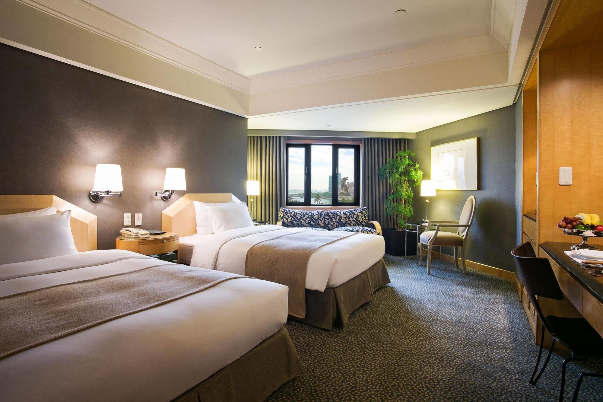 Ambassador Hotel Kaohsiung Exterior photo The photo shows a hotel room featuring two neatly made beds with white bedding and light brown accents. There are bedside lamps on either side of the beds. A large window lets in natural light, and there’s a small sitting area with a couch and a plan