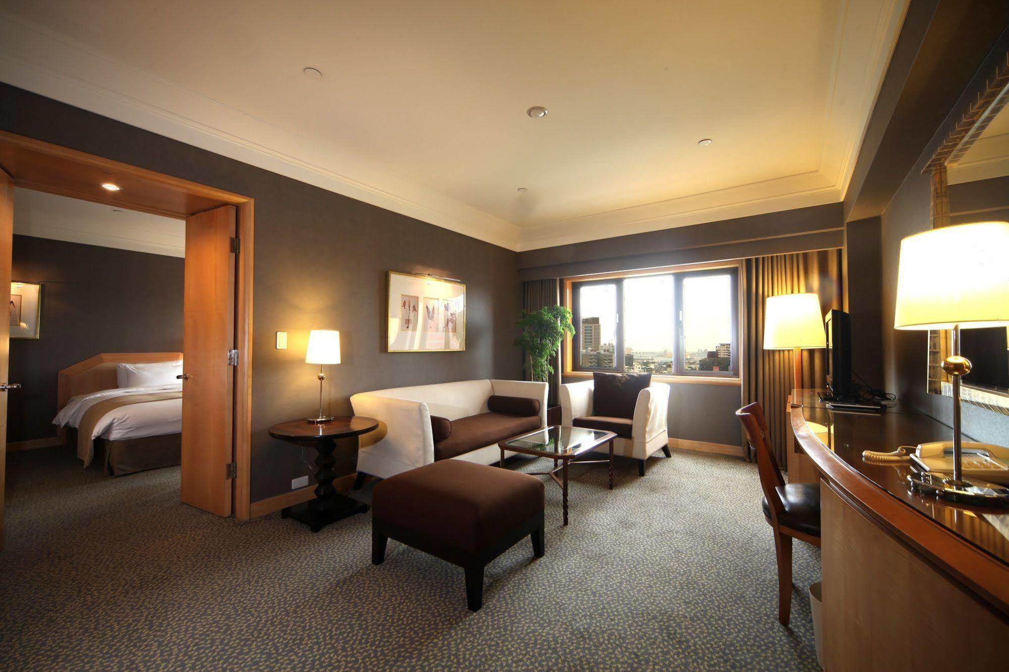 Ambassador Hotel Kaohsiung Exterior photo The photo shows an elegantly furnished hotel room. The space features a comfortable seating area with a white sofa and a dark brown ottoman placed in front of a glass coffee table. There is a small side table with a lamp beside the sofa. A large wind