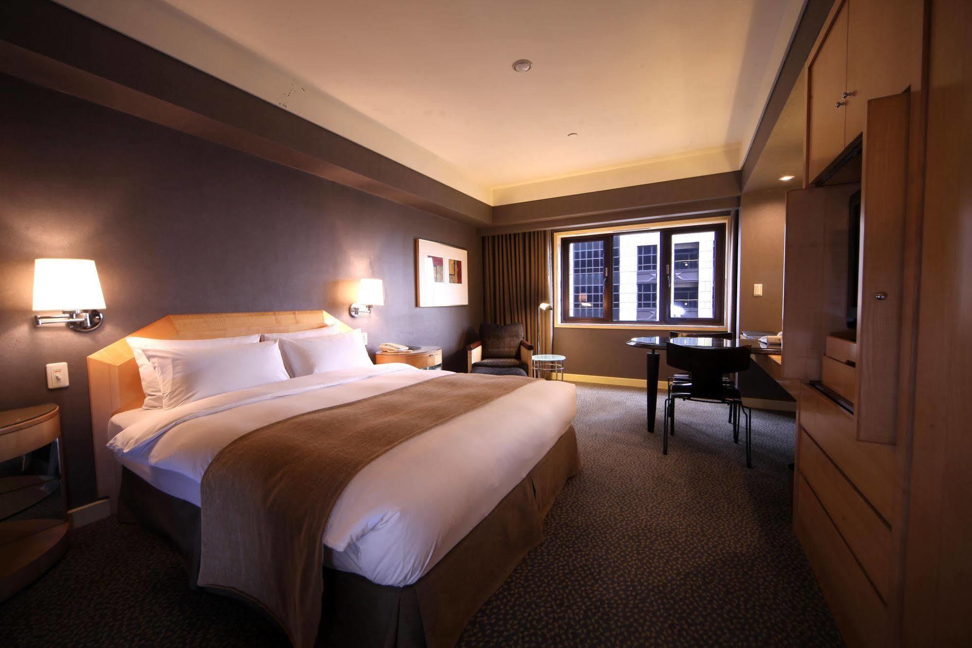 Ambassador Hotel Kaohsiung Exterior photo The photo shows a hotel room featuring a large bed with a white bedspread and a brown accent. There are nightstands on either side of the bed, each with a lamp. A chair is positioned near a window, which offers a view outside. A wooden desk and chair