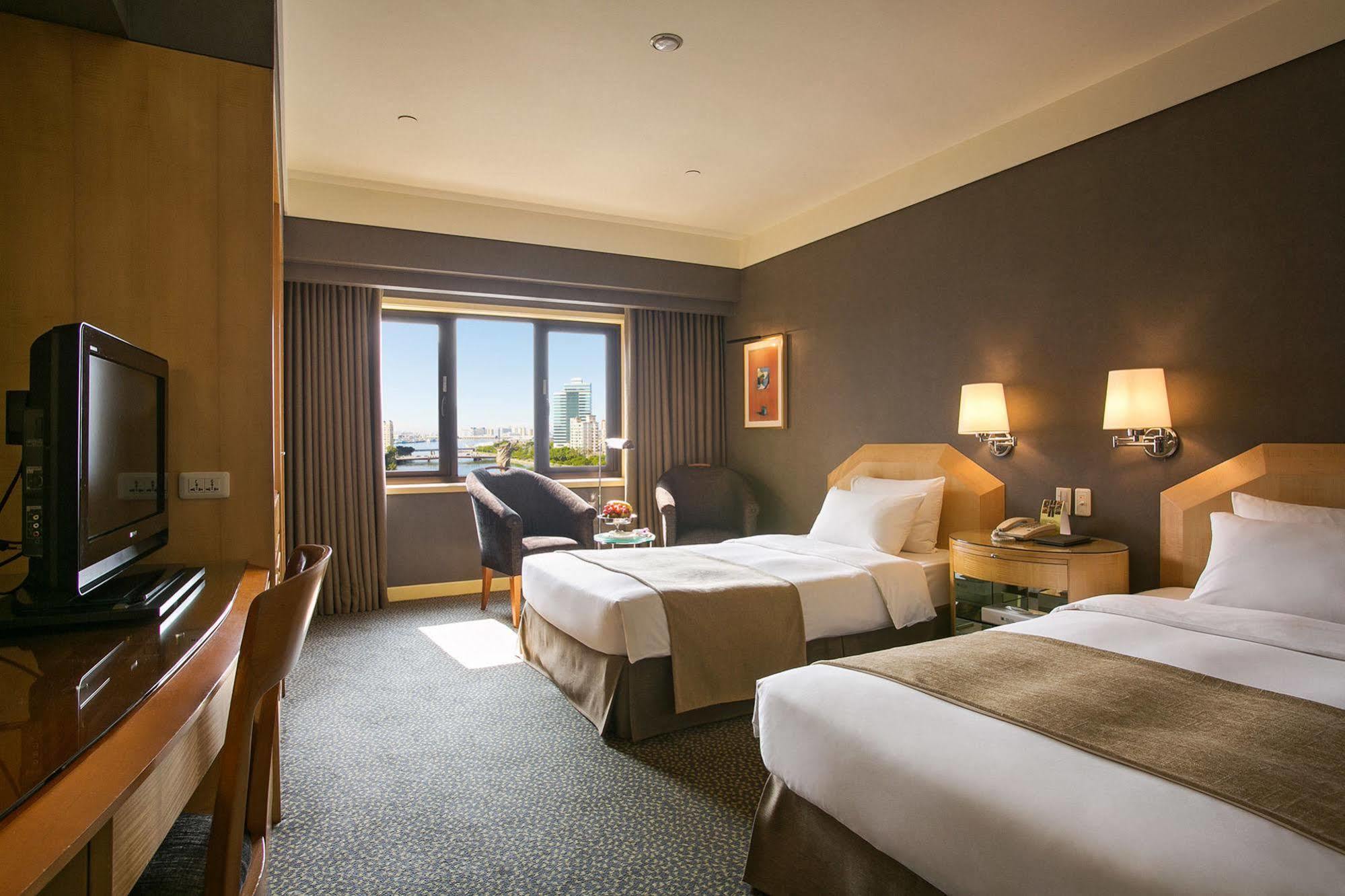 Ambassador Hotel Kaohsiung Exterior photo The photo shows a hotel room with two beds, one of which is a double bed and the other appears to be a single. The room has a modern design with a neutral color palette, featuring wooden furniture, a TV unit, and a small desk and chair. There are two