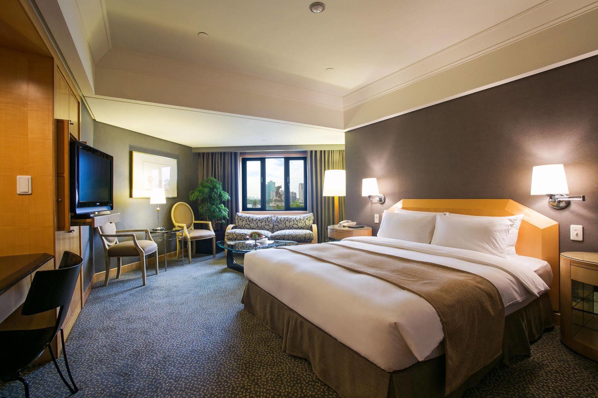Ambassador Hotel Kaohsiung Exterior photo The photo shows a well-appointed hotel room. It features a large bed with white and beige bedding, a comfortable seating area with a sofa and chairs, and a small coffee table. A flat-screen television is mounted on the wall opposite the bed. There ar