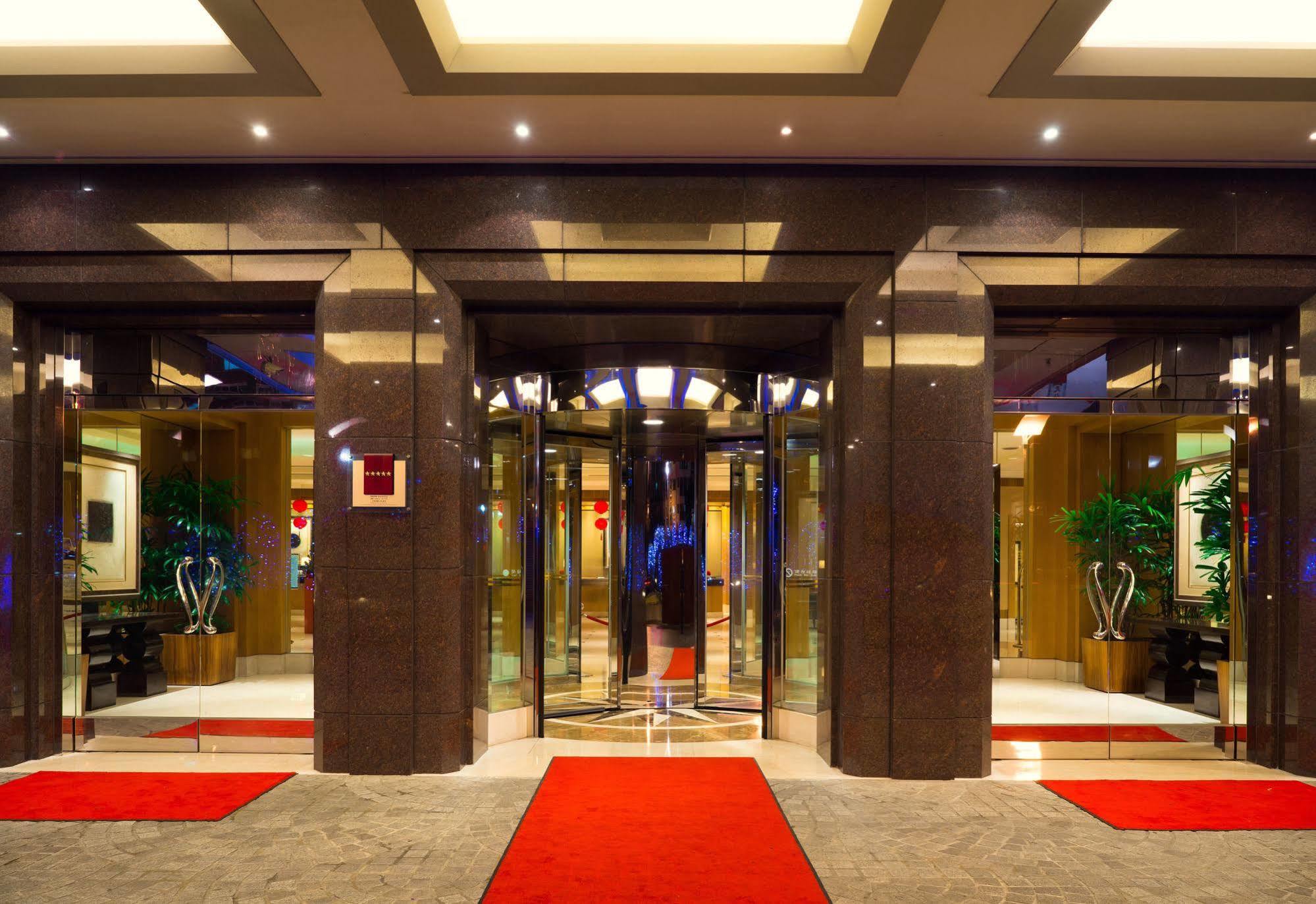 Ambassador Hotel Kaohsiung Exterior photo The photo shows the entrance of a modern hotel or building. It features large glass doors in a sleek frame, creating an inviting entrance. The floor is covered with a plush red carpet leading up to the doors. Inside, there are decorative plants on ei