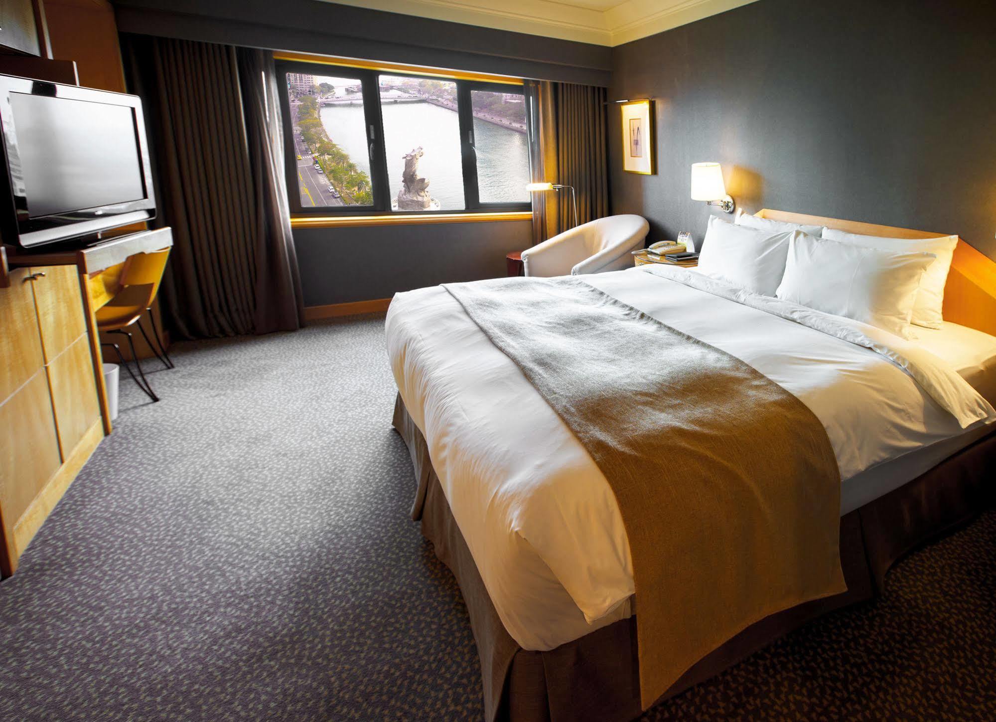 Ambassador Hotel Kaohsiung Exterior photo The photo depicts a modern hotel room featuring a spacious layout. In the center, there is a large bed with crisp white linens and a decorative brown throw blanket. To the side, there is a comfortable armchair and a small table. A television is mount