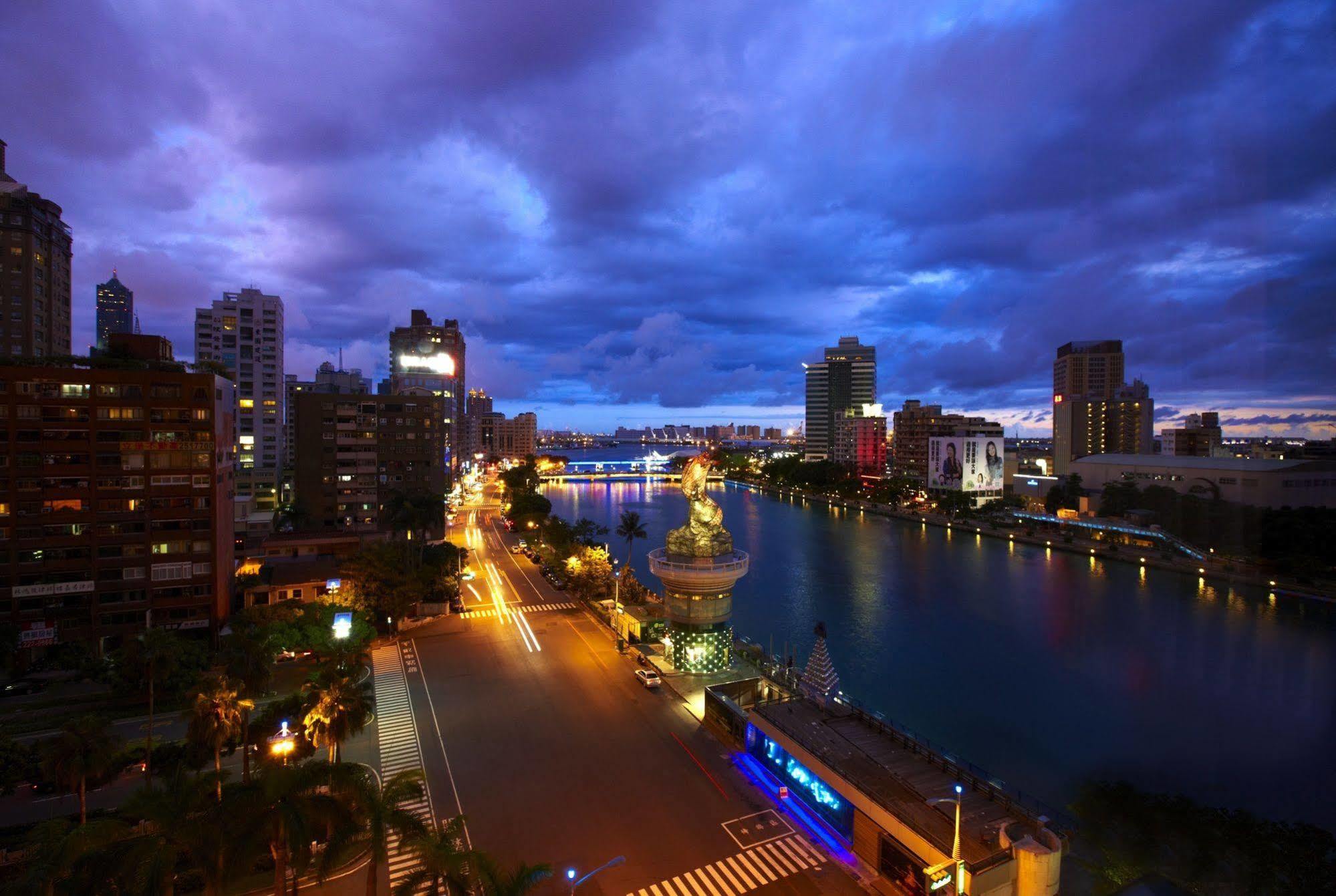 Ambassador Hotel Kaohsiung Exterior photo The photo depicts a cityscape during twilight. On the left, you can see a wide street lined with buildings, and the area is illuminated by streetlights. In the center, there is a body of water, potentially a river or canal, reflecting the lights from