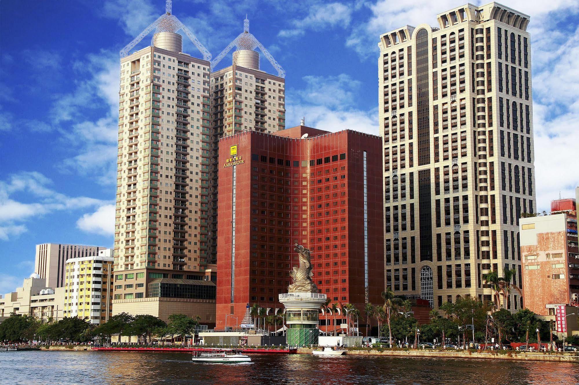 Ambassador Hotel Kaohsiung Exterior photo The photo shows a cityscape featuring several tall buildings, including two prominent skyscrapers with distinctive architectural features. The structures are situated near a body of water, suggesting a waterfront location. The sky is blue with some c