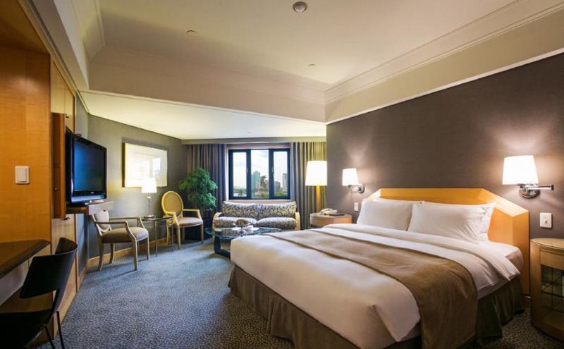 Ambassador Hotel Kaohsiung Exterior photo The photo shows a hotel room that features a large bed with white linens and a light brown throw blanket. There are two bedside lamps on either side of the bed. In the corner, there is a cozy seating area with a patterned sofa and two chairs. A telev