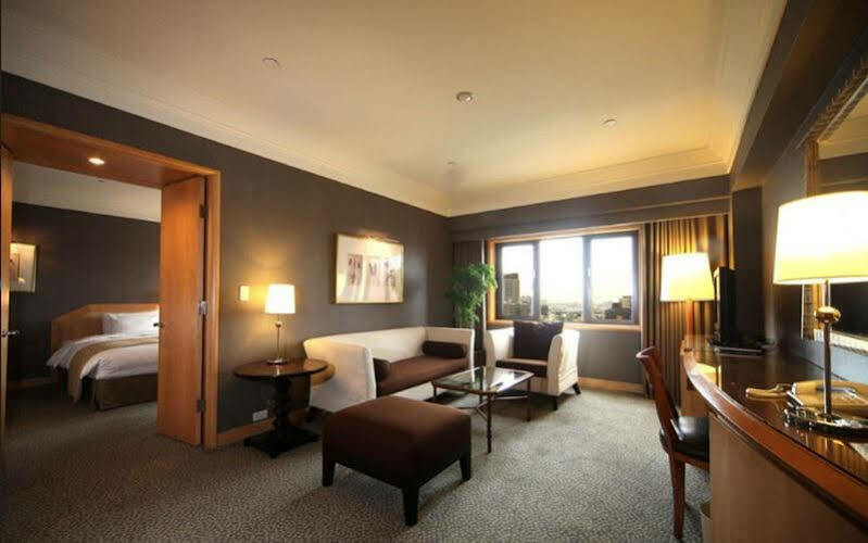 Ambassador Hotel Kaohsiung Exterior photo The photo shows a stylish hotel room with modern décor. There is a comfortable seating area featuring a white sofa and brown ottoman, along with a glass coffee table. A side table with a lamp is positioned next to the sofa. The room has a large windo